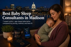 Mother holding a sleeping baby by a window with a nighttime Madison skyline; desk items include a candle, a Madison mug, and a phone, for a guide to Best Baby Sleep Consultants in Madison (2026).