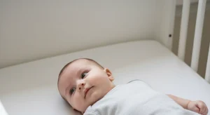 Baby lying on a white blanket in a crib, head tilted and looking toward the camera.