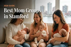 Cover image for 2026 guide: three smiling moms with babies on a sofa by a window overlooking a city, captioned 'Best Mom Groups in Stamford'.