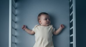 Sleeping baby on their back in a cream onesie in a blue crib.