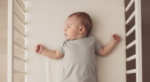 Baby sleeping on a light crib mattress, wearing a gray onesie, arms resting at sides within a white crib frame