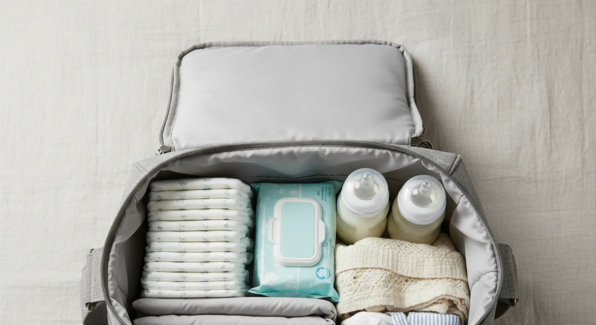 Open diaper bag on a bed with diapers, wipes, bottles, and baby clothes neatly arranged