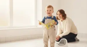 Cheerful fully clothed toddler boy standing beside a small potty chair in a bright bathroom holding a picture book