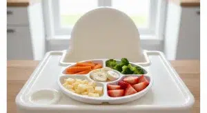 Balanced toddler meal on a divided high-chair tray with quartered grapes, steamed veggie sticks, cheese cubes, and soft pasta in a bright kitchen.