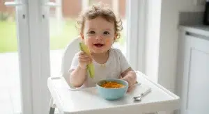 Baby in a high chair holding a strip of avocado next to a bowl of sweet potato puree