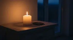 Single lit candle beside a rosary on a wooden table in a quiet room at night