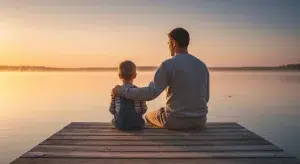 Father and his young son sitting side by side on a dock at sunrise, both fully in frame from behind