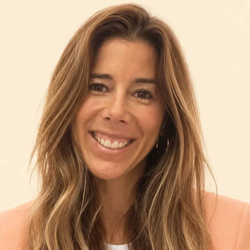 Smiling woman with long wavy light brown hair wearing a peach blazer, looking at the camera.