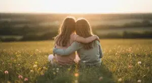 Two young sisters sitting close together on a sunny meadow hill, seen from behind with their arms around each other