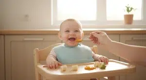 Baby sitting upright in a high chair being spoon-fed orange puree while starting solids