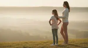 Young girl standing confidently on a grassy hilltop at sunrise with her parent beside her, both looking out at the view