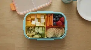 A colorful toddler lunch bento box with cheese, soft carrots, cucumber, halved berries, pasta, and crackers.