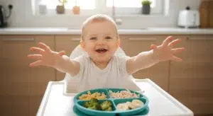 Toddler Meals for Picky Eaters 50 Easy Ideas That Get Eaten | Betteroo Toddler in a high chair reaching for a divided plate of pasta, broccoli, chicken, and banana