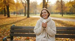 Smiling woman in a cream sweater sits on a park bench holding a cup of coffee among autumn leaves and sunlight.