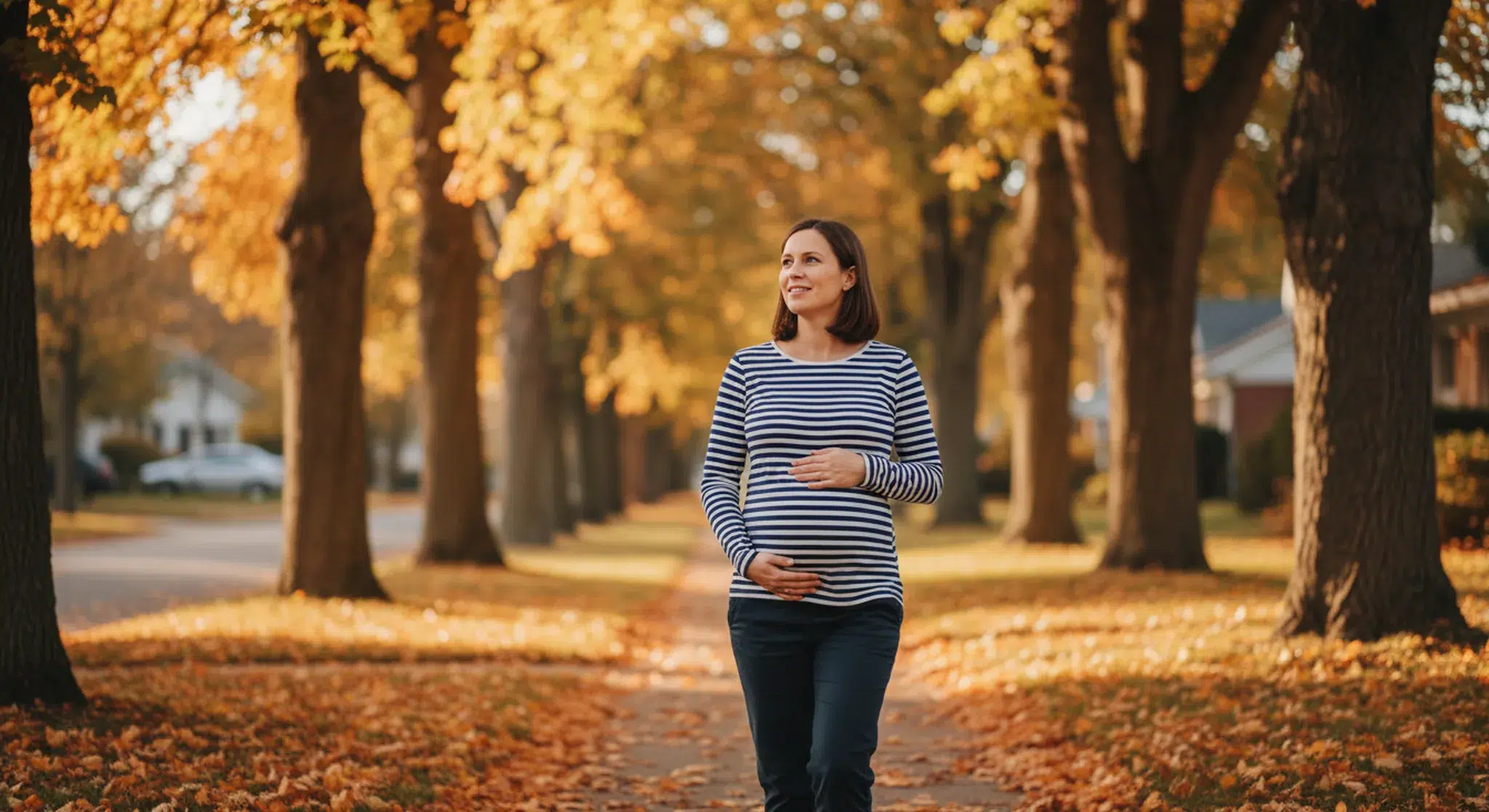 Pregnant woman in a blue-and-white striped long-sleeve top walking along a tree-lined autumn street with orange leaves on the ground.