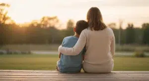 Mother and young son sitting together on a wooden porch at sunset, seen from behind, her arm around him