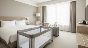 A bright hotel room with an empty mesh play yard holding only a fitted sheet beside a neatly made bed and a closed suitcase on the floor