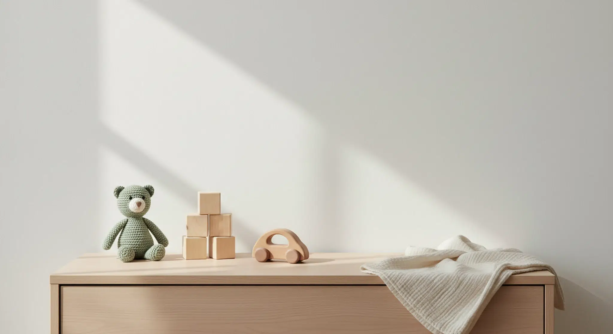 A knitted teddy bear, plain wooden blocks and a wooden toy car on a nursery dresser in soft morning light