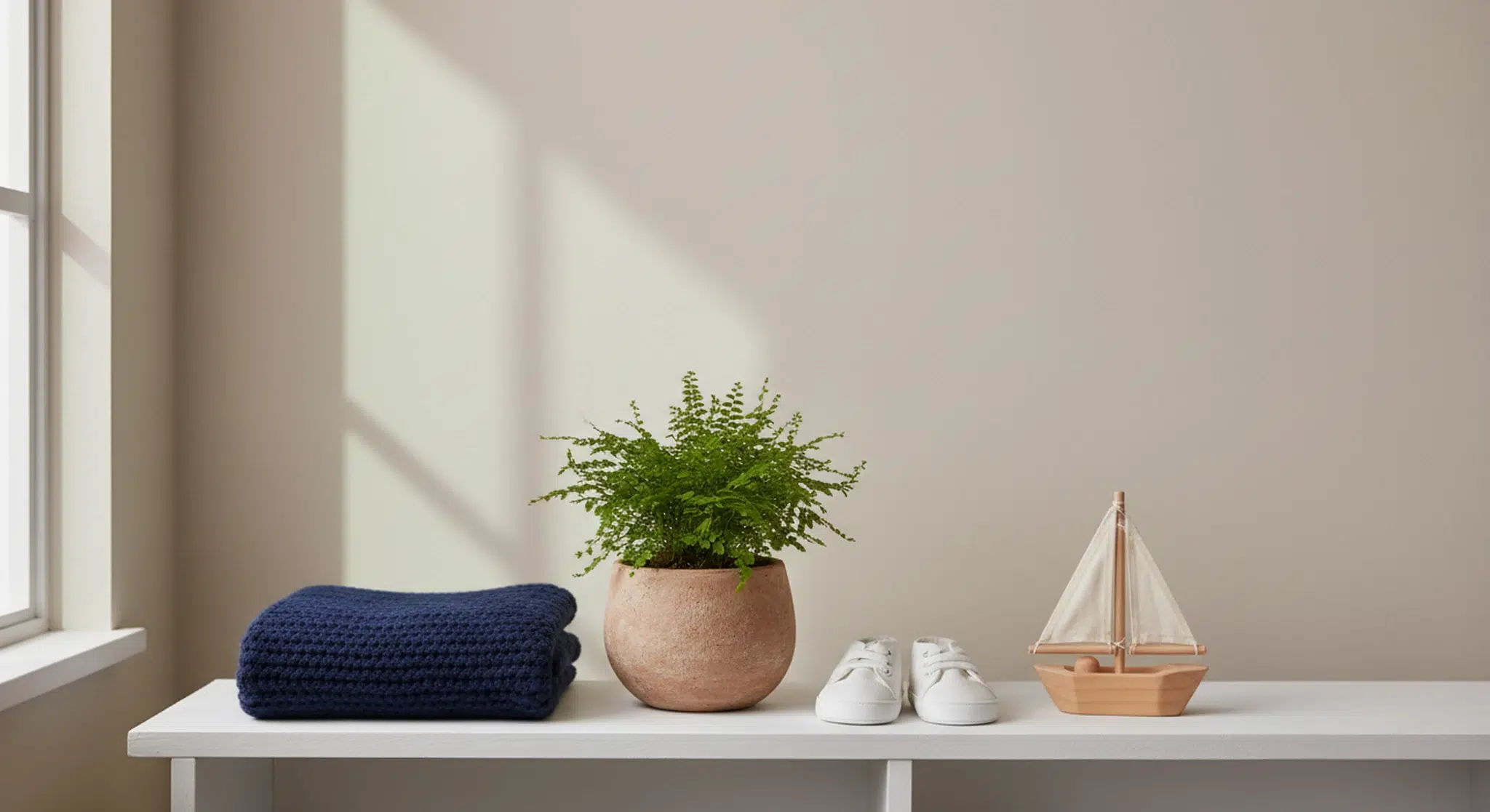 Small potted fern, folded navy knit baby blanket, tiny canvas sneakers and a wooden toy boat on a white shelf