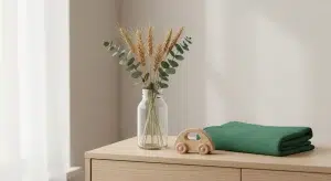 Glass vase with dried wheat and eucalyptus on a light wood dresser, beside a neatly folded green blanket and a wooden toy car.