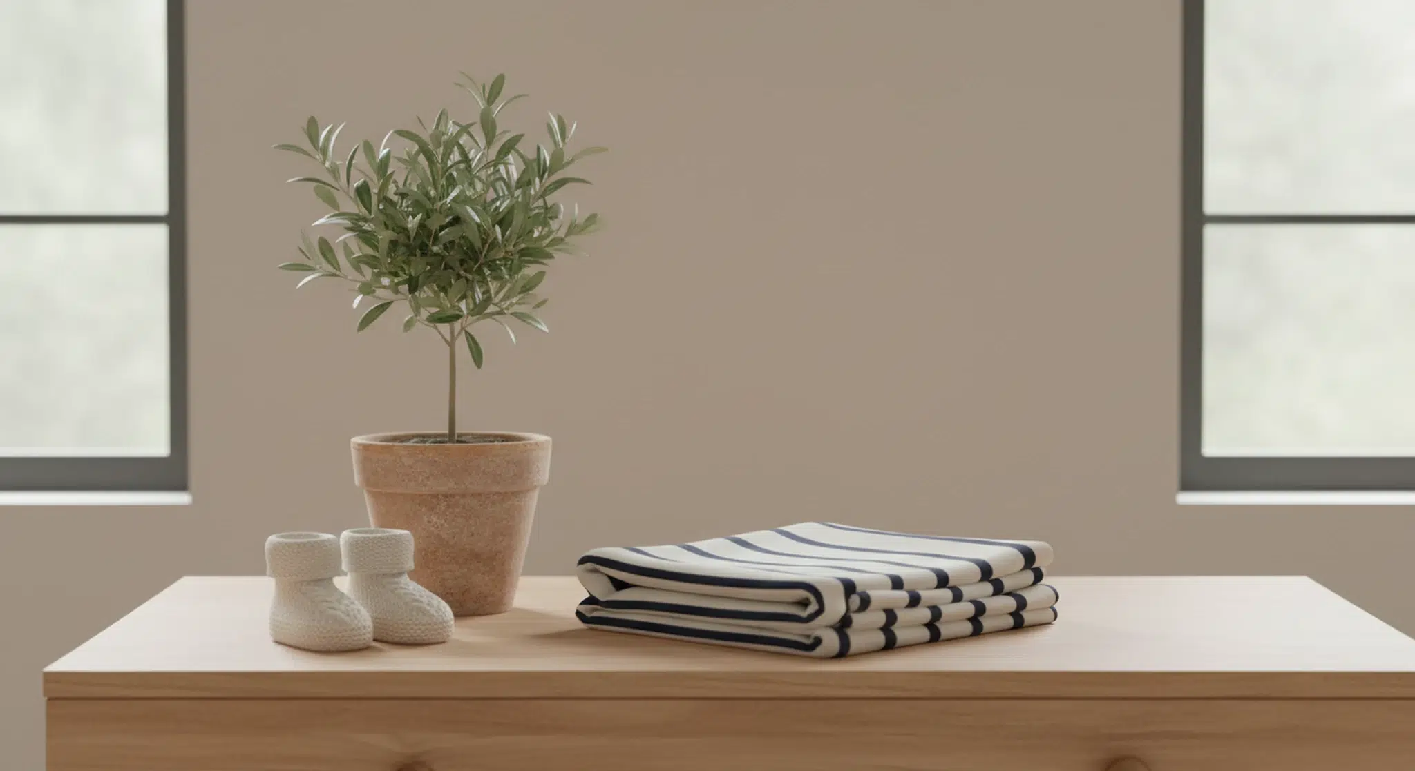 Small potted olive tree, folded navy and white striped baby blanket and tiny knit booties on a light wooden dresser top