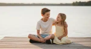 Two children sit on a wooden dock by a calm lake, smiling and embracing each other.