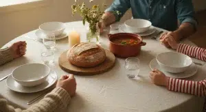 A family gathered at a simple dinner table with rustic bread, vegetable soup, and a lit candle in warm evening light