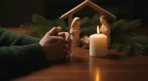 Hands folded in prayer beside a lit candle and a small wooden nativity on a table with pine branches