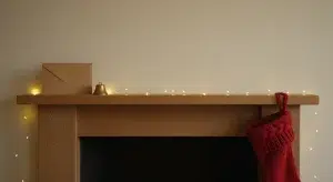 A red knit stocking, a blank kraft-paper envelope and a brass jingle bell on a wooden mantel with warm fairy lights