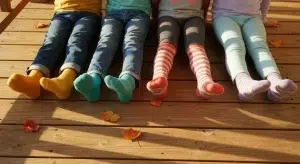 Four children sitting side by side on a wooden porch with their legs stretched out, showing brightly colored socks in warm sunlight
