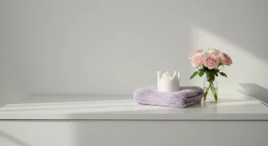 Pink roses in a glass vase placed on a lavender knitted cloth on a white shelf, with a small white crown beside them.