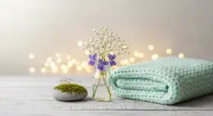 Glass jar with white baby's breath and purple flowers, next to a moss ball and a mint knitted blanket, under warm bokeh lights.