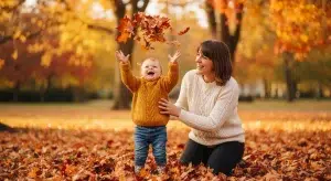 Toddler in a mustard sweater tossing autumn leaves in a sunlit park while a smiling parent kneels beside them