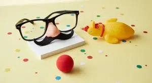 Novelty disguise glasses with a nose and mustache on blank index cards beside a rubber chicken, a red clown nose and paper confetti on a pale yellow table