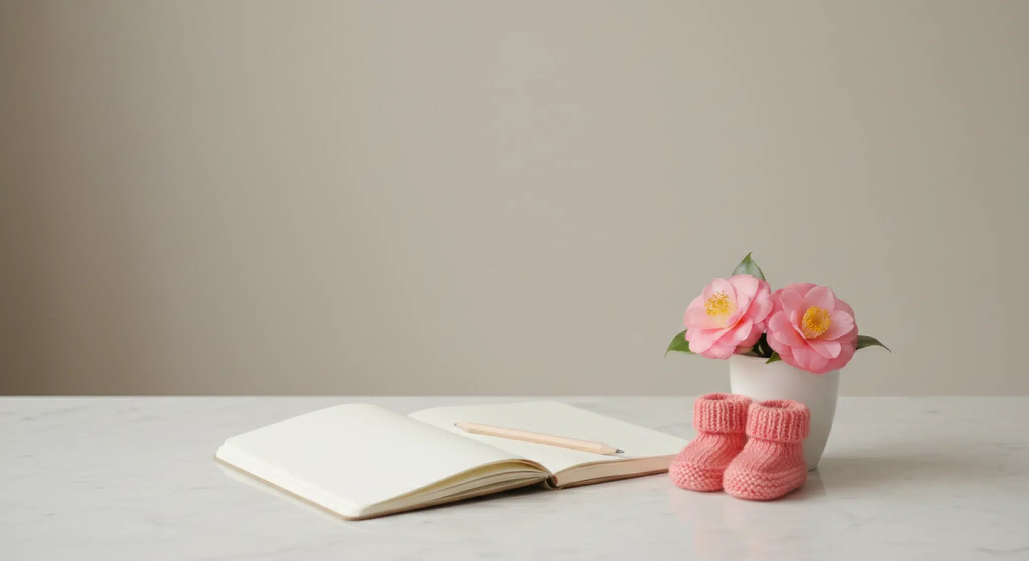 Blank cream notebook, pencil, a small cup of pink camellia blooms and coral-pink knitted baby booties on a light marble tabletop