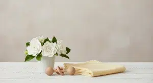 A small white ceramic cup of gardenias, a folded pale yellow muslin swaddle and a wooden rattle on a white-washed wooden table in soft daylight