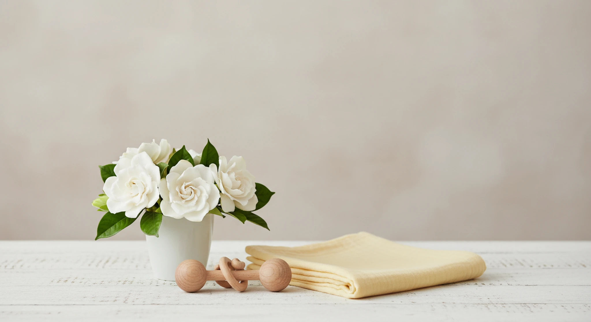 A small white ceramic cup of gardenias, a folded pale yellow muslin swaddle and a wooden rattle on a white-washed wooden table in soft daylight