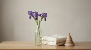 A few stems of purple iris in a clear glass bottle, a folded cream knit baby blanket and a small wooden ring stacker on a light oak table in soft morning light