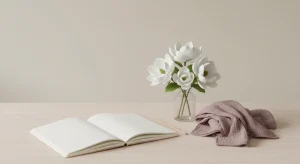 Blank cream notebook, pencil, a small glass of white magnolia-like blossoms and a folded dusty-mauve muslin baby swaddle on a pale birch tabletop
