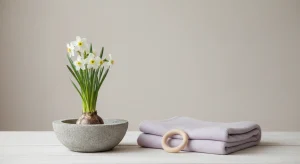 White narcissus flowers in a low stone bowl beside a folded pale lavender baby blanket and a wooden teething ring on a pale wooden table