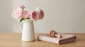 Girl Names That Start with R 133 R Names with Origins and Meanings | Betteroo Pink roses in a white ceramic pitcher on a wooden table, with a pink cloth and wooden teething ring beside them.