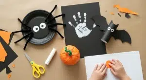 Overhead view of a kraft paper covered table with a paper plate spider, a handprint ghost, a paper bag pumpkin and a toilet roll bat, and a child's hands pressing a sticker