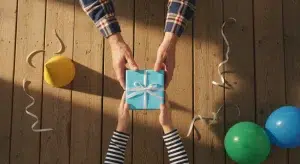 An older man's hands passing a small gift wrapped in plain blue paper to a young boy's hands, seen from above on a wooden porch
