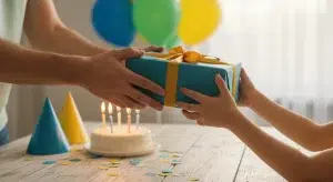 Adult hands passing a gift wrapped in blue paper to a young boy's hands across a table with a birthday cake and candles