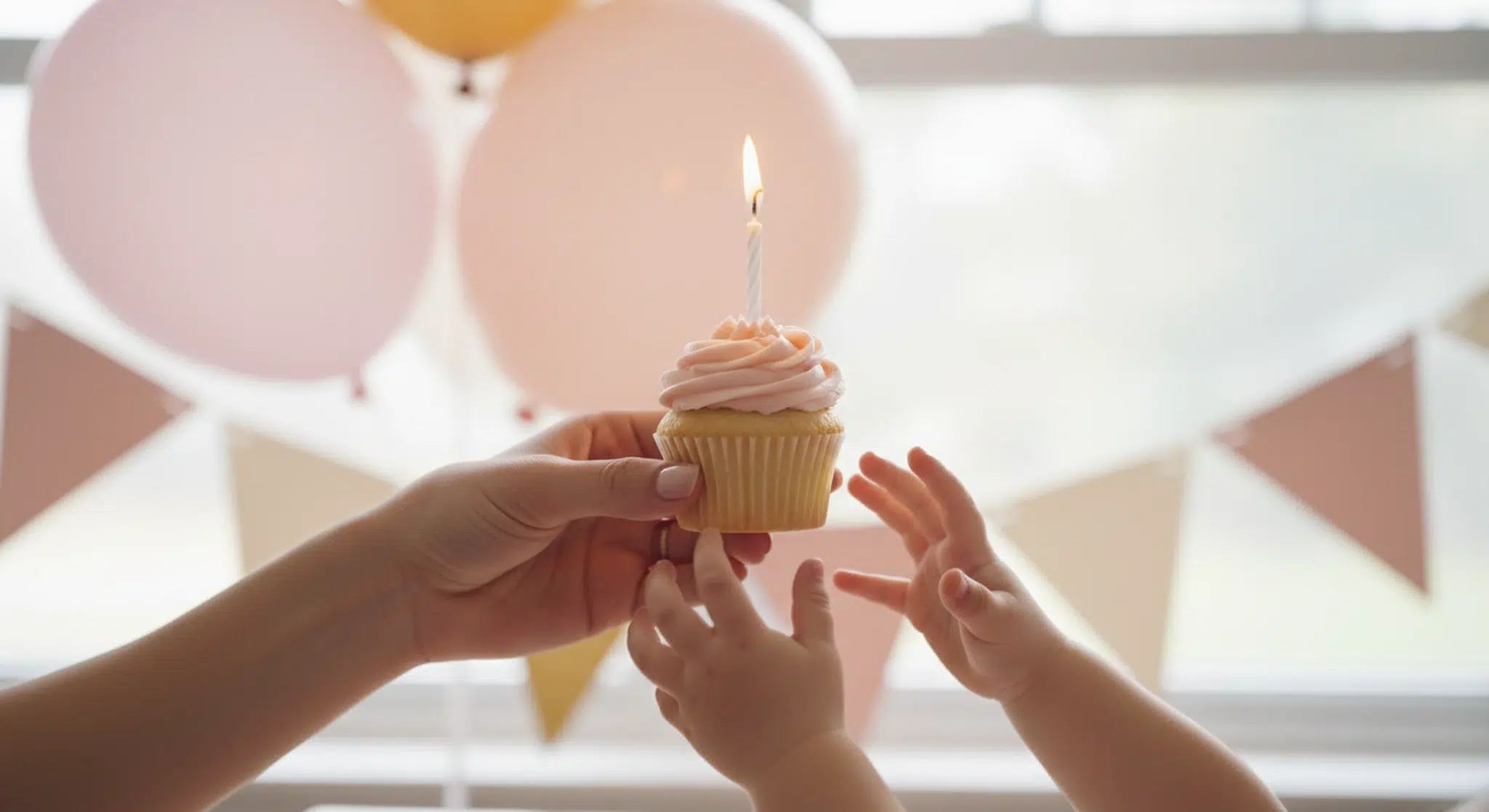 Adult hands holding out a cupcake with one lit candle toward a small child's hands, with pink and gold balloons blurred behind