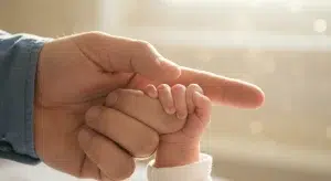 A baby's tiny hand wrapped around a father's finger, his plain gold wedding band catching soft morning light