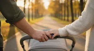 A couple's hands with wedding bands resting together on the handle of a baby stroller on a tree-lined path at golden hour