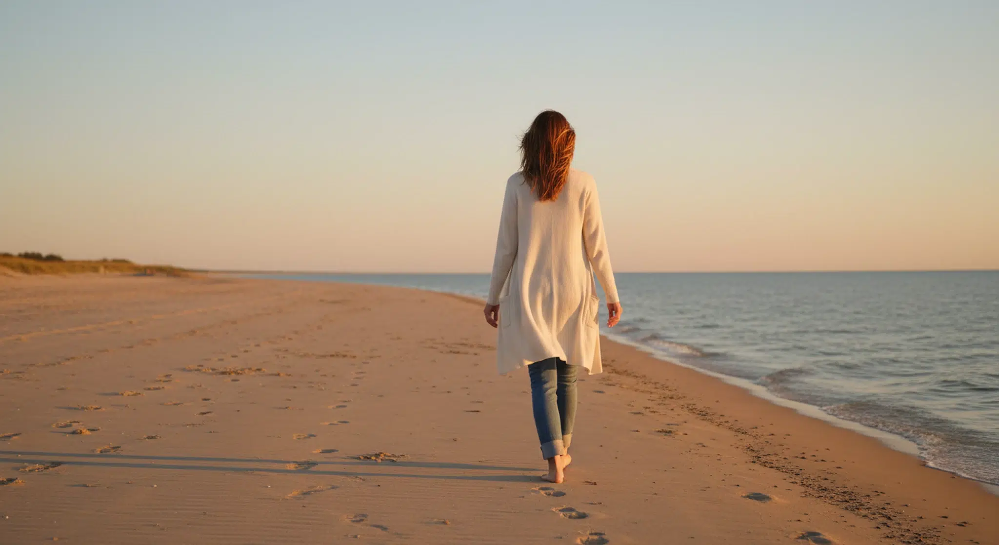 Woman walking alone along a quiet beach at golden hour, seen from behind