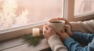 A parent and child's hands holding a warm mug together by a frosty window at sunrise