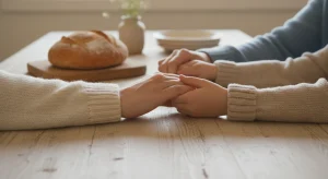 Family members holding hands across a wooden dinner table beside a loaf of bread and a vase of wildflowers in soft window light